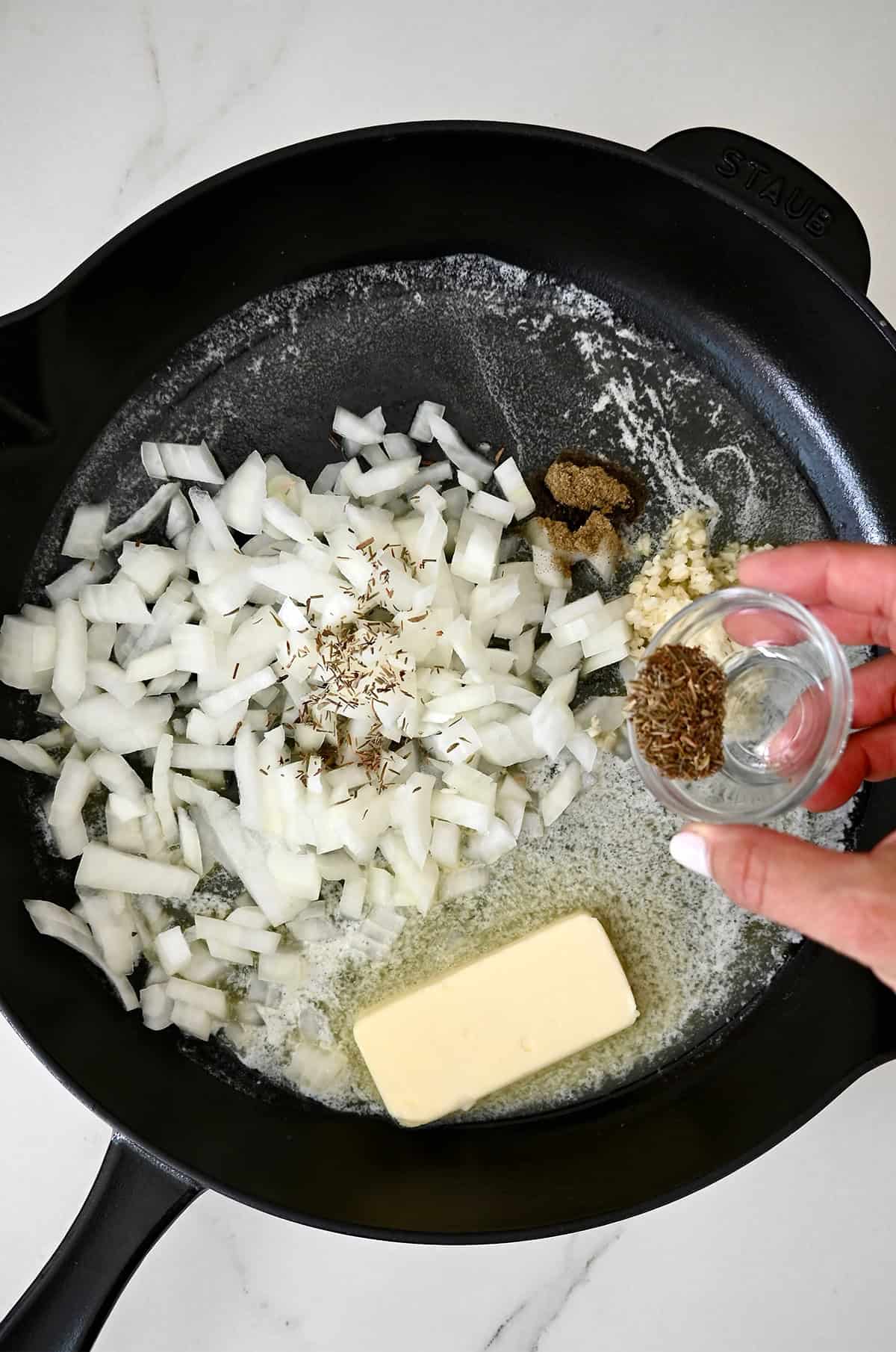 Dried herbs are being added to a cast-iron skillet with diced onions, garlic, and half a stick of butter.