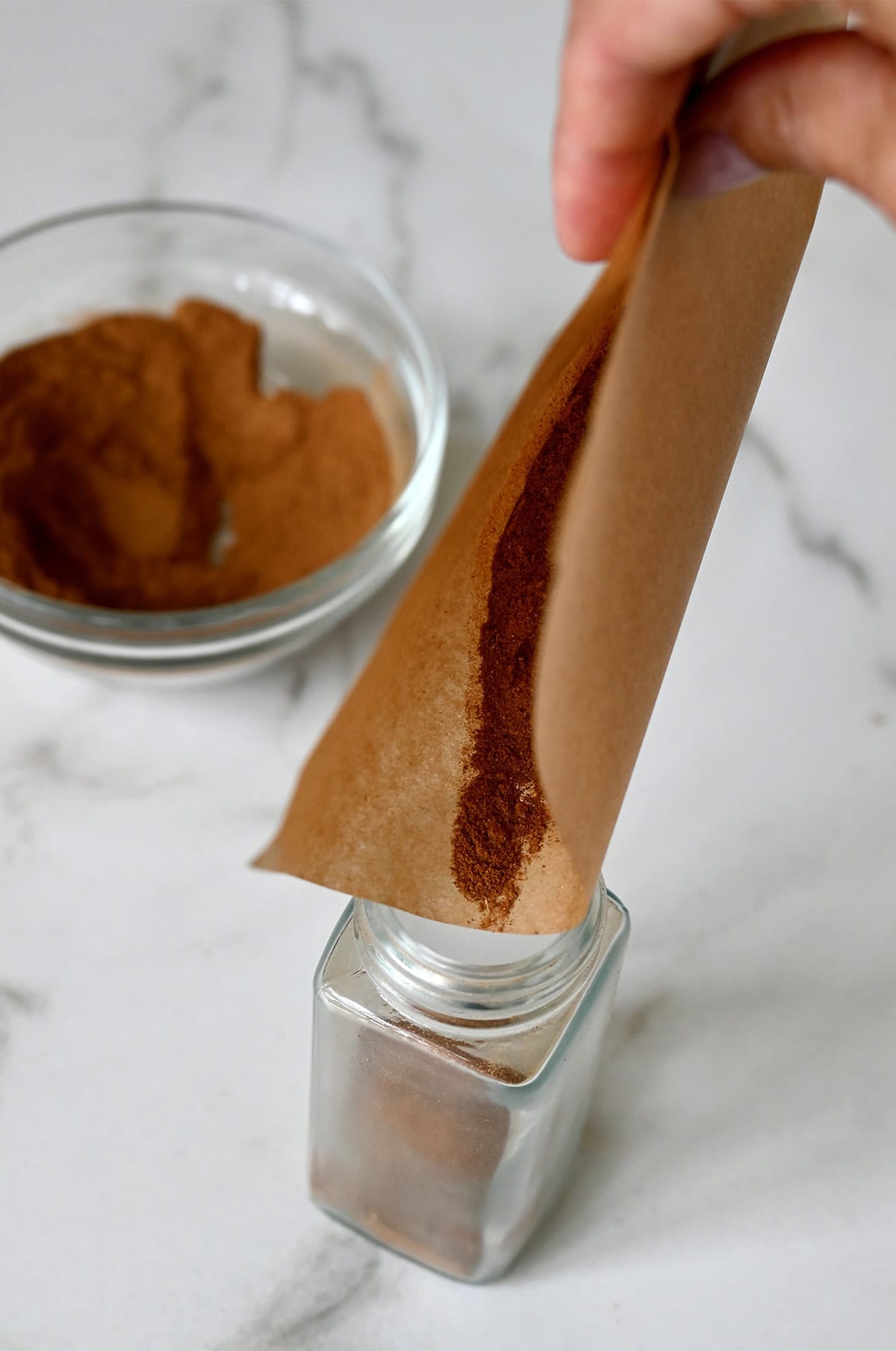 Using a piece of parchment paper as a funnel to transfer homemade pumpkin spice into a glass jar.
