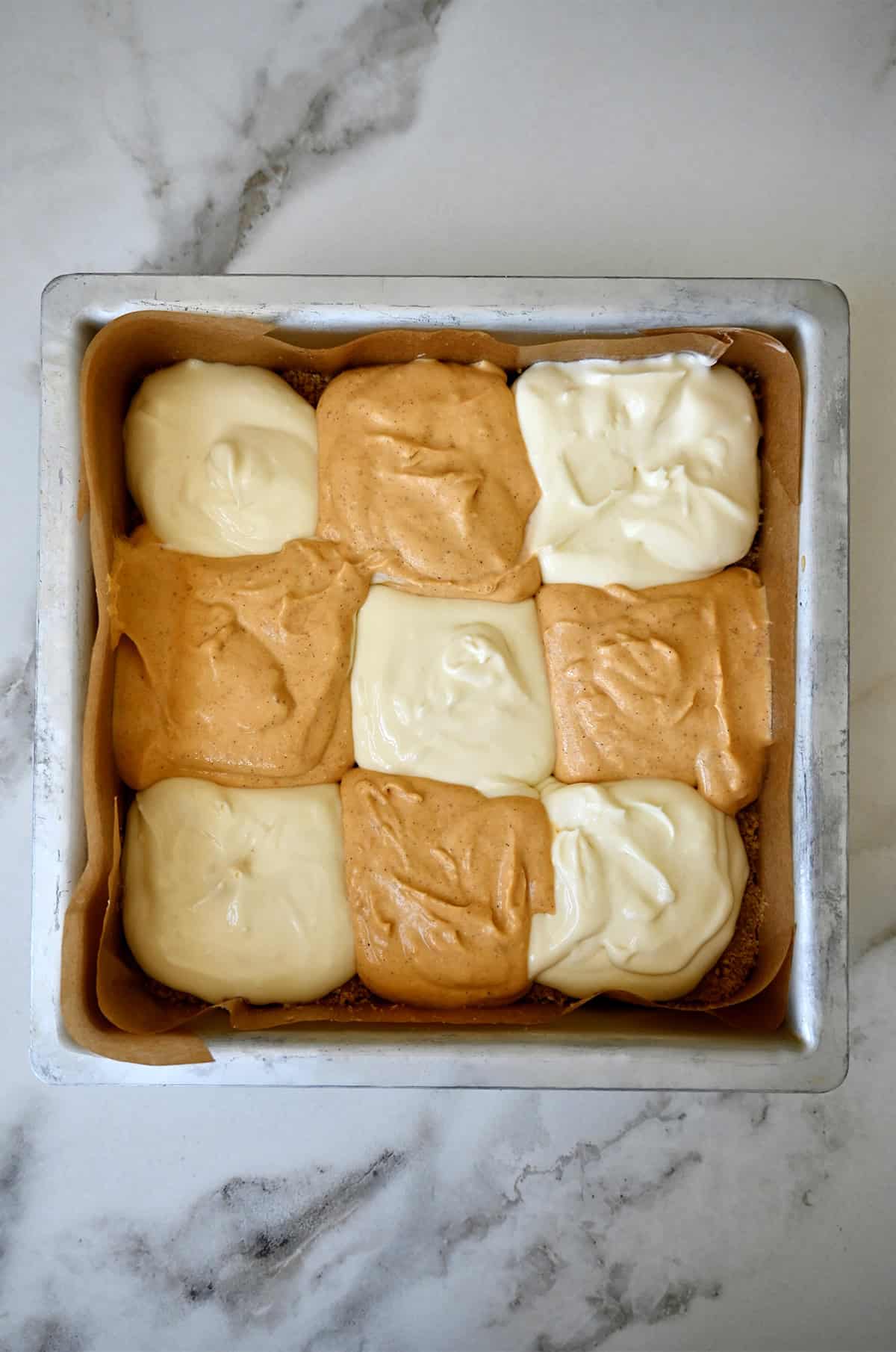 Checkerboard pattern of cheesecake and pumpkin pie batters in a square baking pan lined with parchment paper.