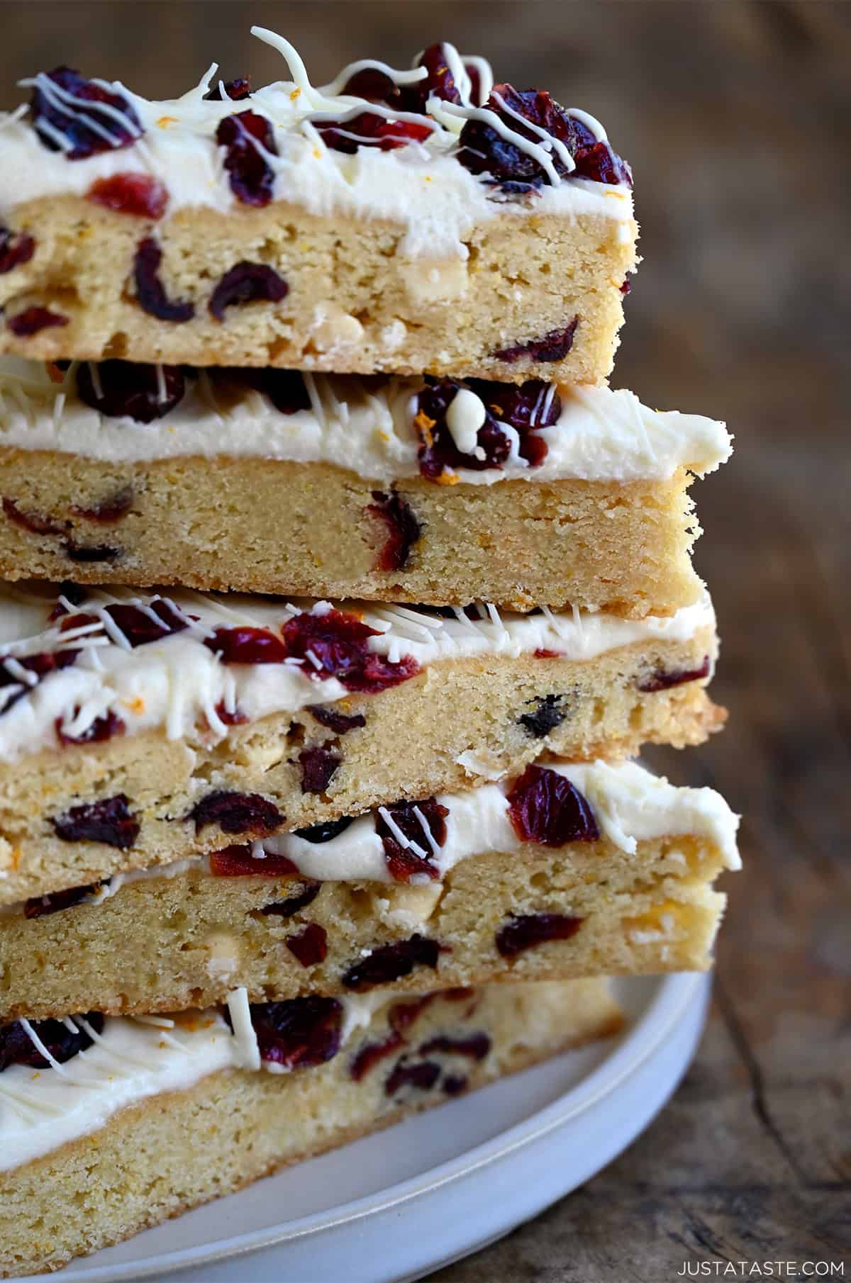 A tall stack of homemade cranberry bliss bars studded with dried cranberries and white chocolate chips, topped with a tangy cream cheese frosting and orange zest.