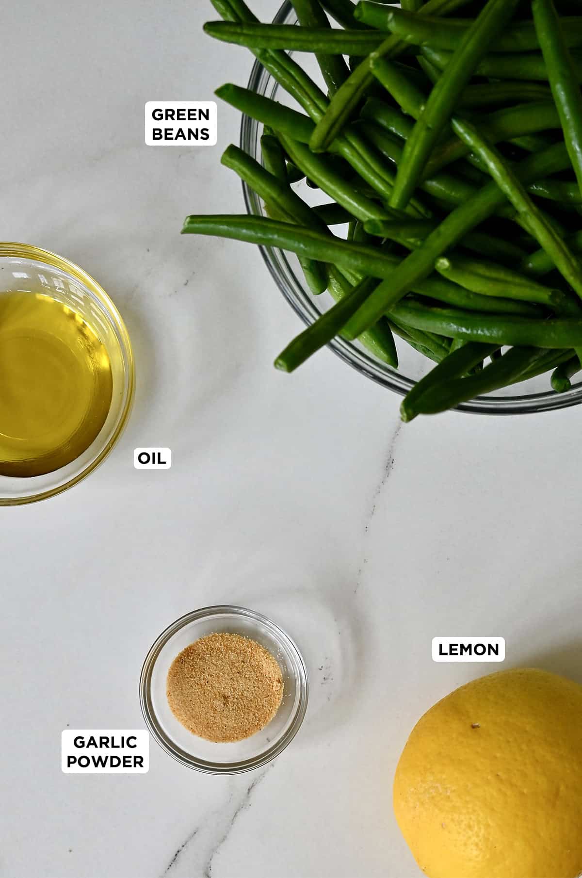 Green beans in a glass bowl next to a lemon and smaller bowls containing olive oil and garlic powder.