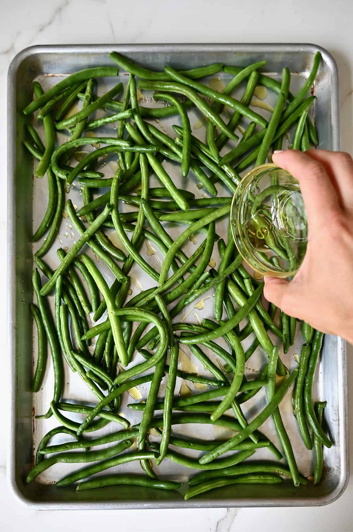 Pouring olive oil from a small bowl atop fresh green beans on a baking sheet.