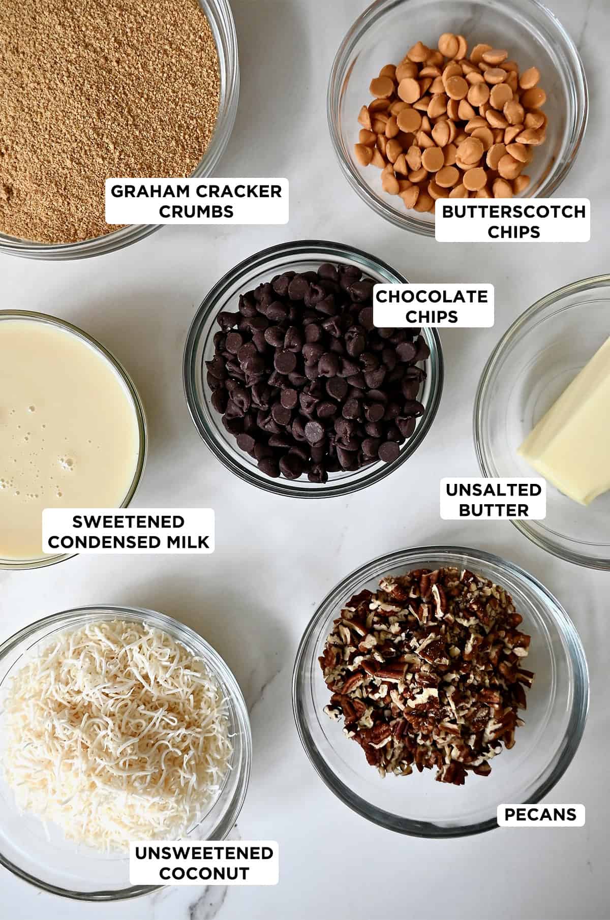 Glass bowls containing ingredients to make magic cookie bars, including crushed graham crackers, melted butter, butterscotch chips, sweetened condensed milk, flaked coconut, chopped pecans and semisweet chocolate chips.