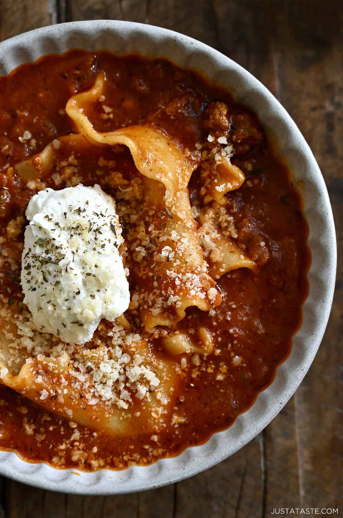 A bowl containing ground beef lasagna soup topped with grated Parmesan, a dollop of ricotta cheese and freshly cracked black pepper.