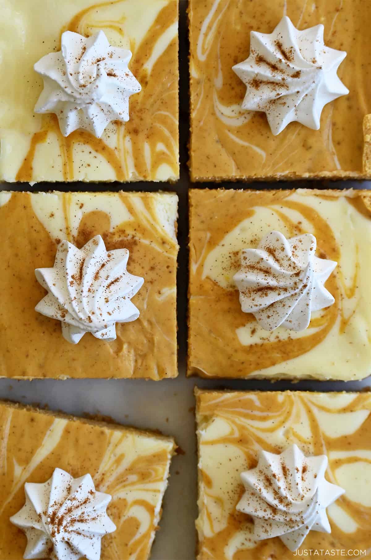 Creamy pumpkin cheesecake bars with swirls of plain cheesecake and pumpkin pie, and a graham cracker crust. Each bar is topped with a dollop of whipped cream and a sprinkle of pumpkin pie spice.