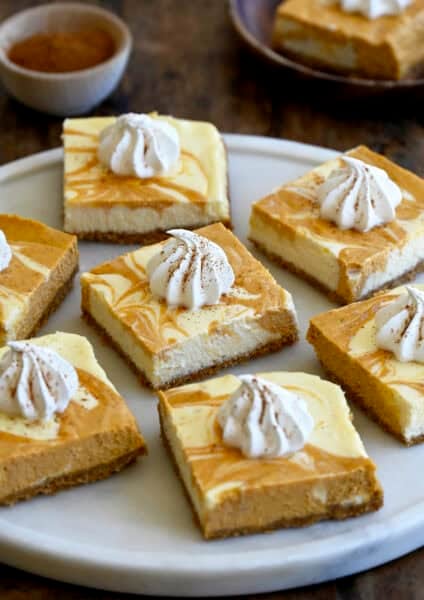 Pumpkin cheesecake bars topped with a dollop of whipped cream and a sprinkle of nutmeg on a round serving plate.