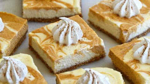 Pumpkin cheesecake bars topped with a dollop of whipped cream and a sprinkle of nutmeg on a round serving plate.