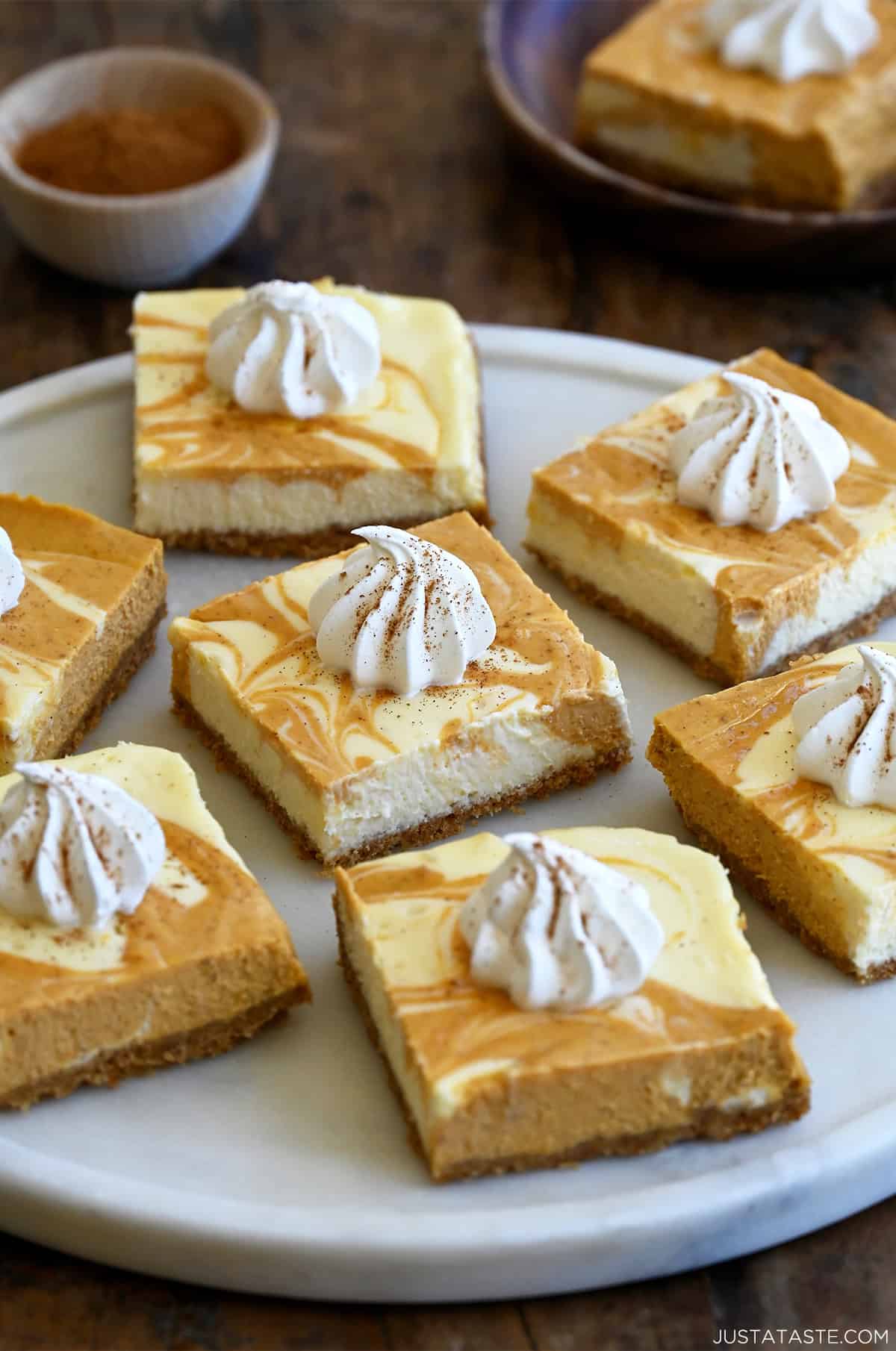 Pumpkin cheesecake bars topped with a dollop of whipped cream and a sprinkle of nutmeg on a round serving plate.