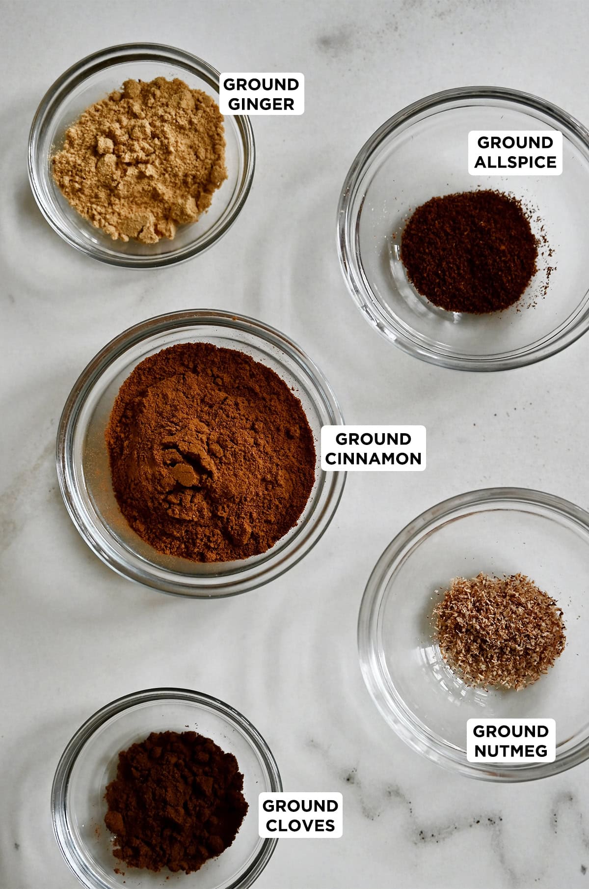 Glass bowls containing the spices that make up pumpkin pie spice, including ground cinnamon, allspice, nutmeg, ground cloves and ground ginger.