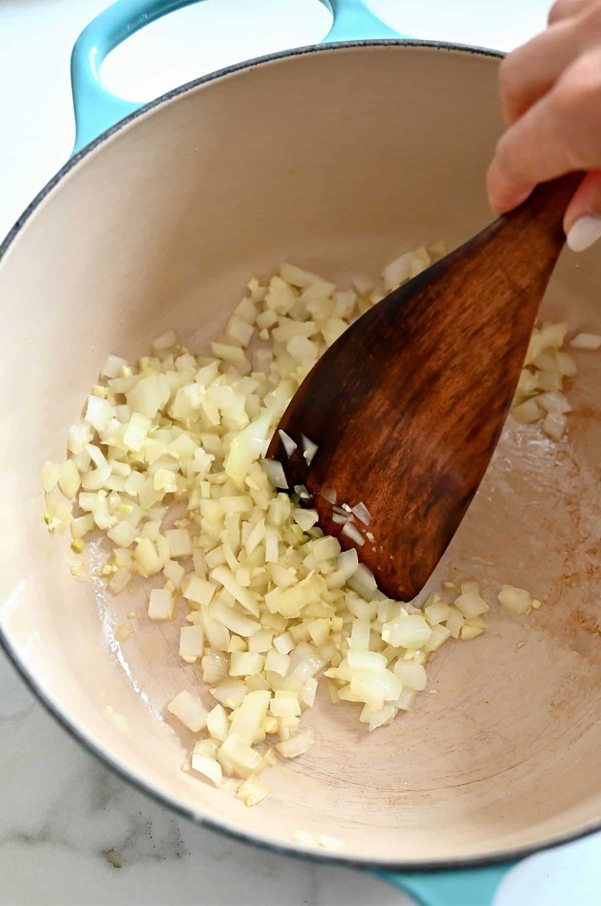 Sauteed onions and garlic in a Dutch oven are being pushed around with a wooden spoon.