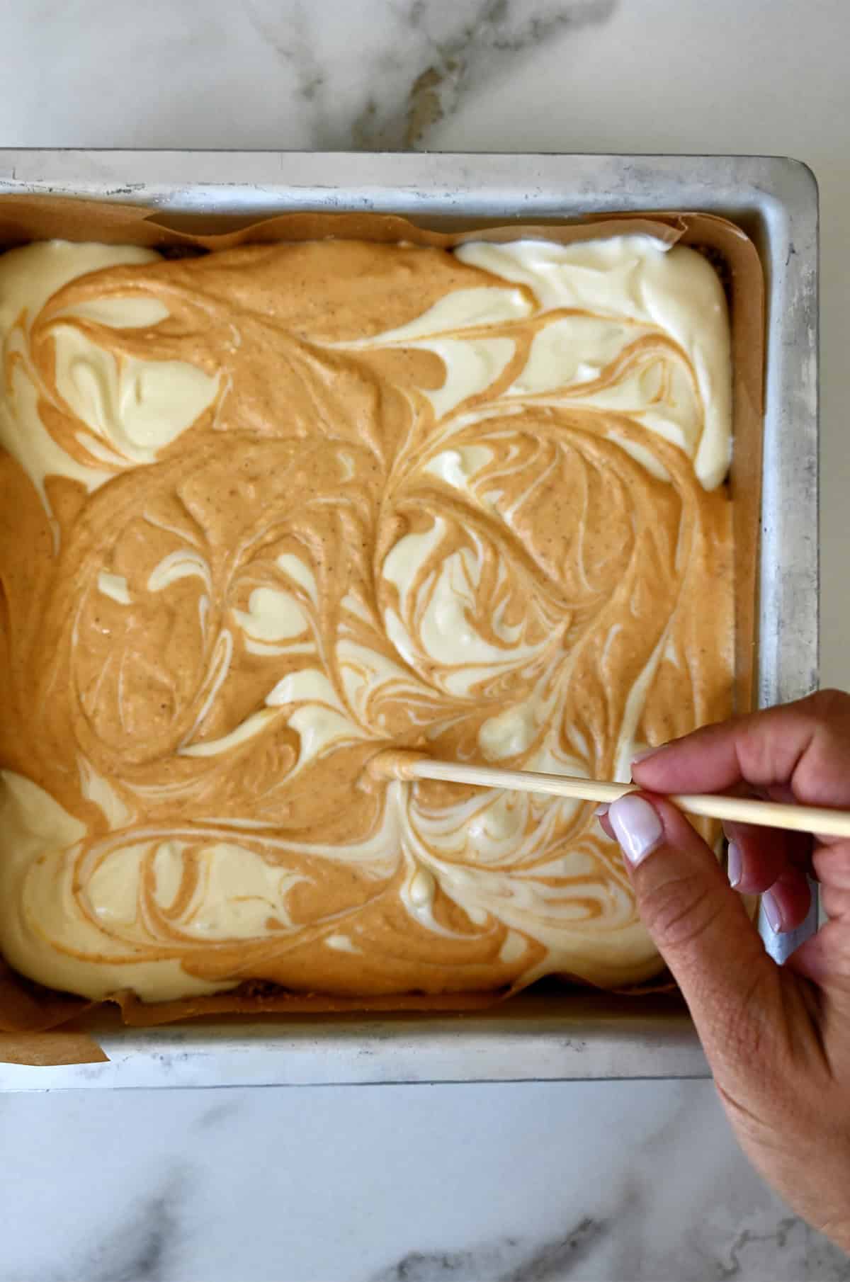 A hand holding a wooden skewer swirls together pumpkin and cheesecake batters in a parchment paper-lined pan.