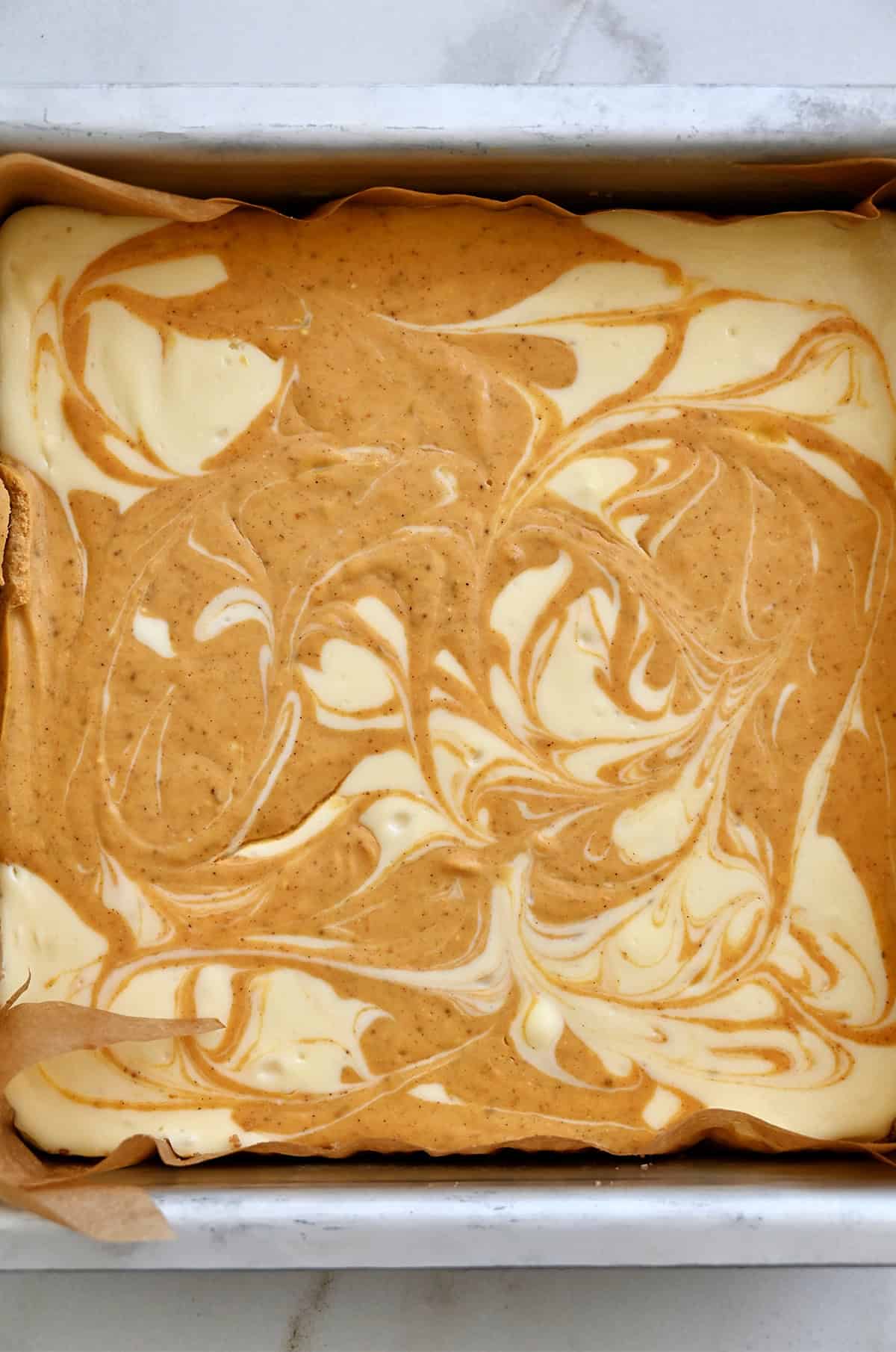 Swirled pumpkin cheesecake bars in a square parchment paper-lined baking pan.