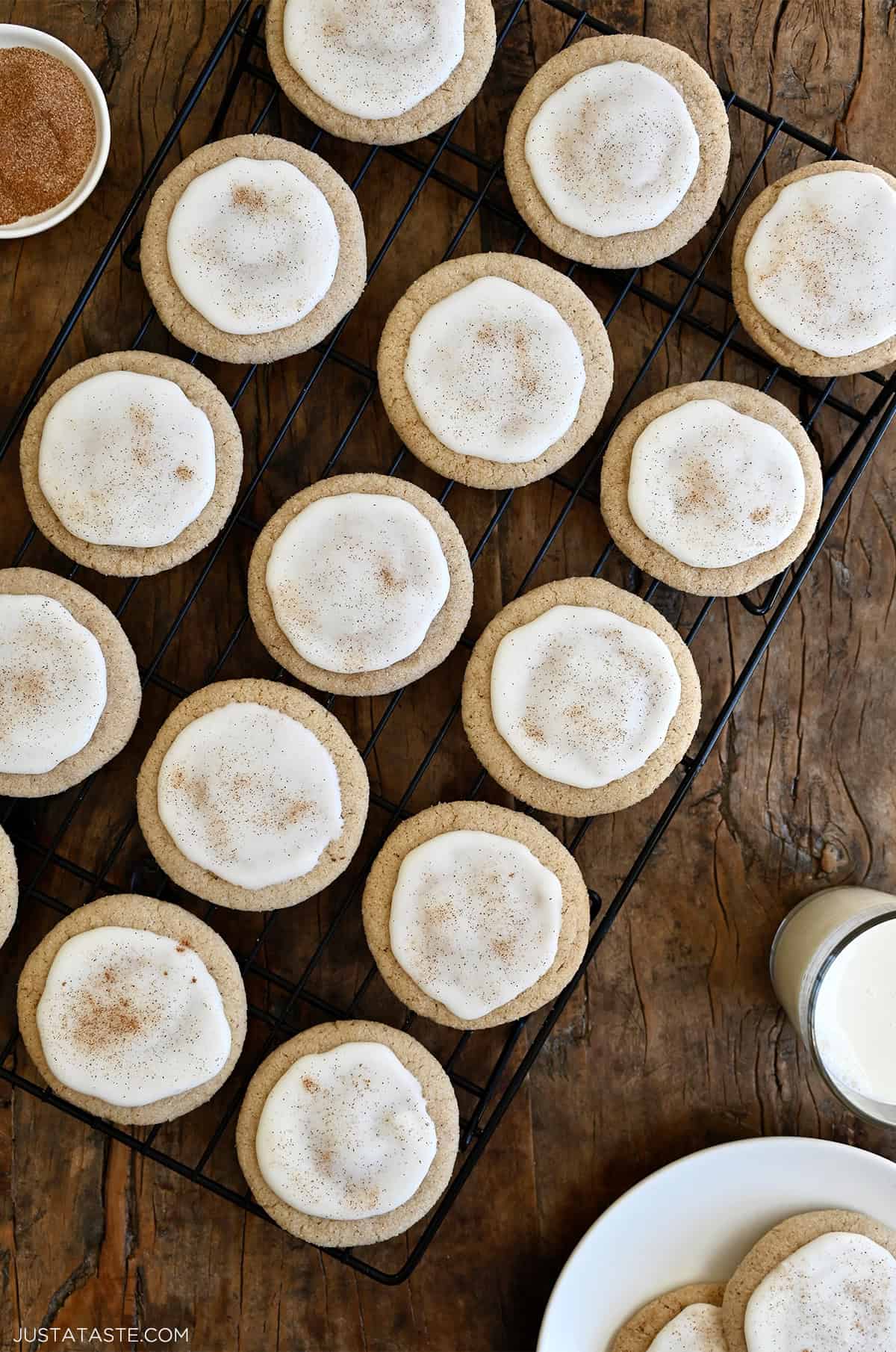 Chai-spiced sugar cookies topped with icing and a sprinkle of cinnamon cooling on a wire rack next to a glass of milk. 