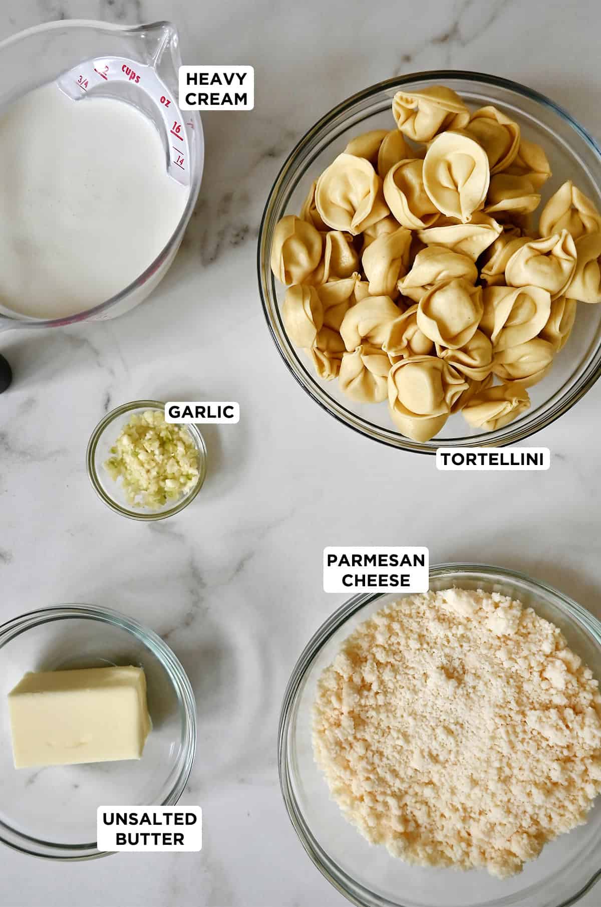 The five ingredients to make tortellini Alfredo in glass bowls: refrigerated tortellini, heavy cream, minced garlic, grated Parmesan cheese and butter.