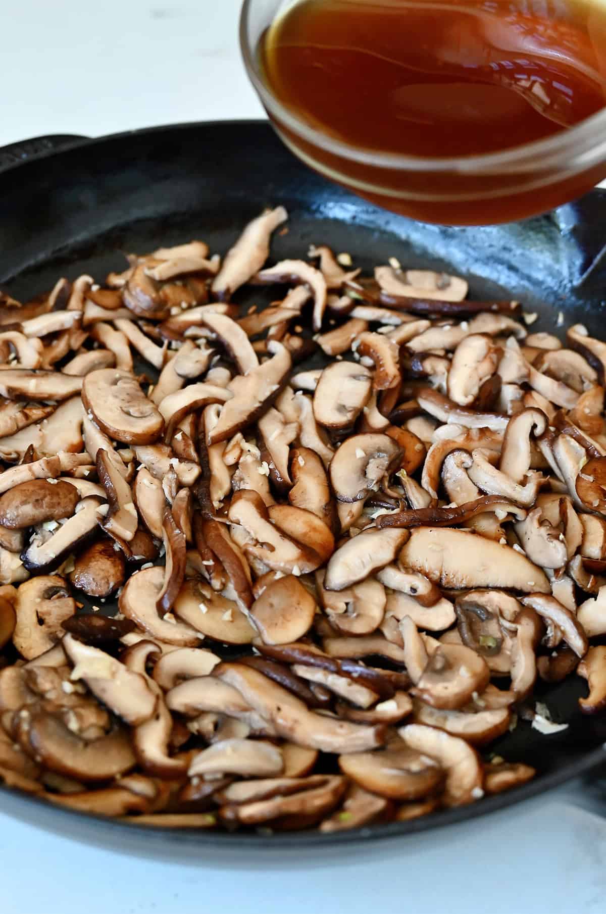 Adding beef stock to a large skillet with sautéed mushrooms and garlic.