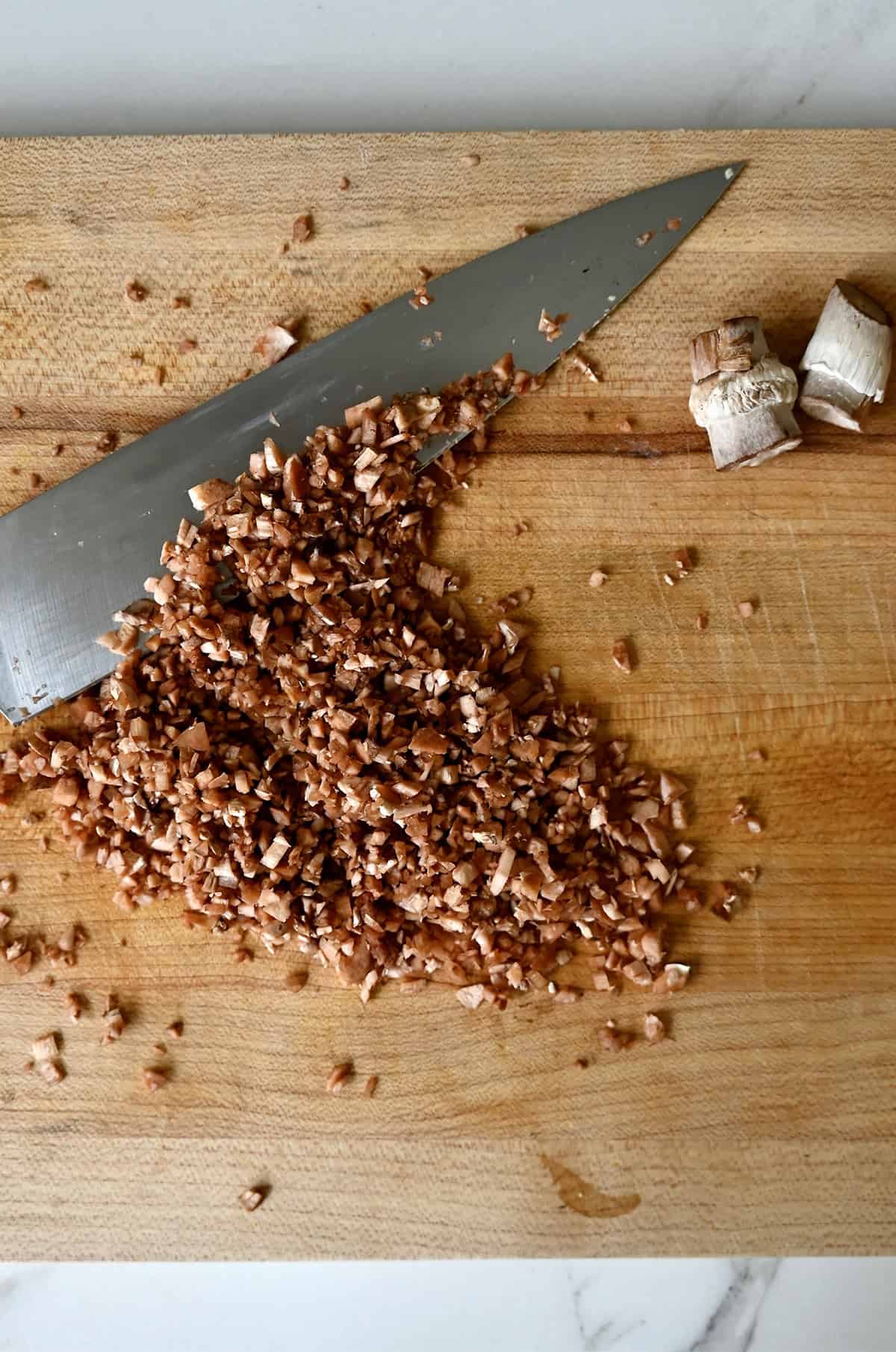 Finely chopped mushroom stems on a cutting board with a sharp knife.