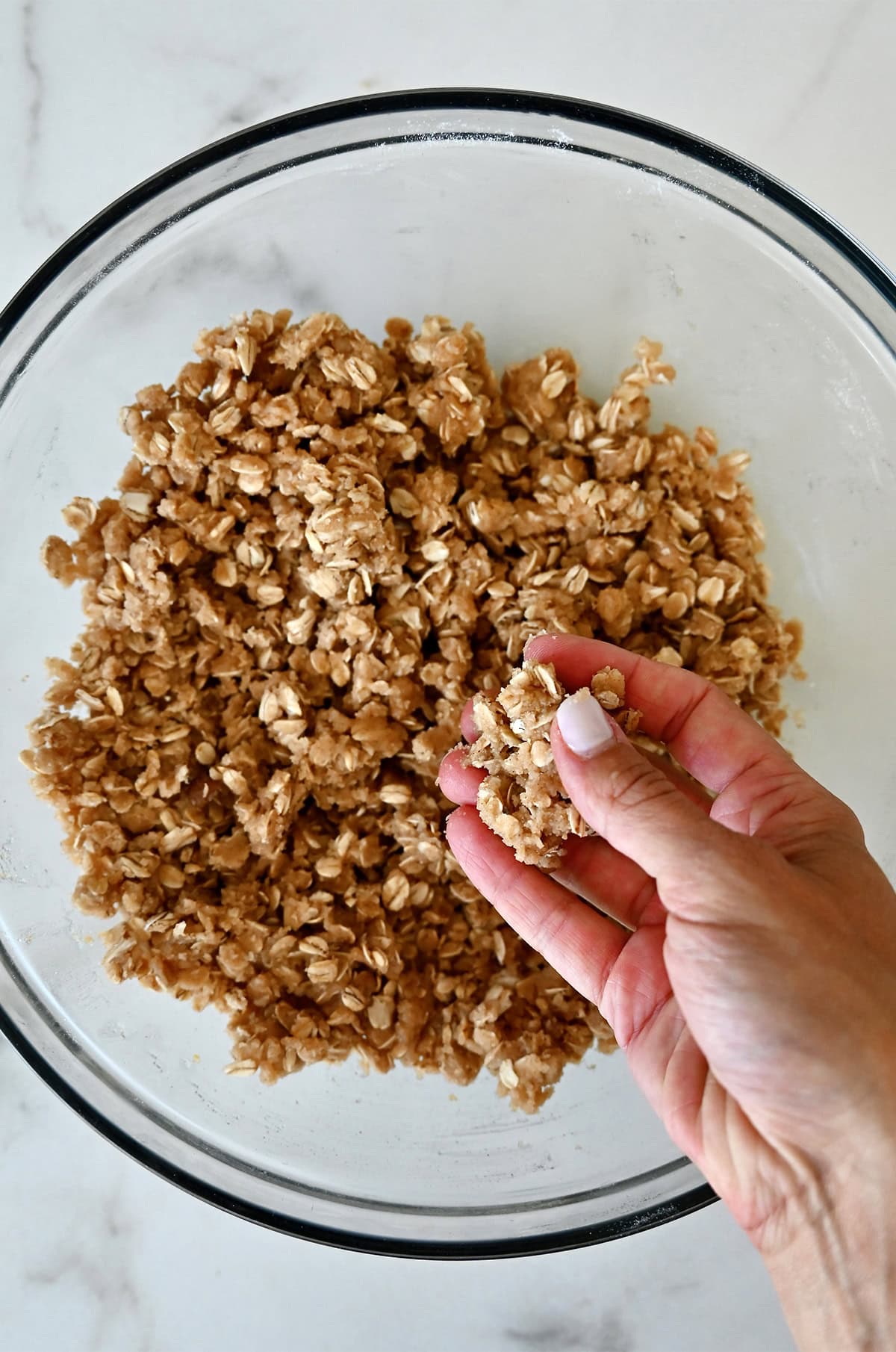 A hand holds a chunky, cookie-like crumble mixture above a bowl with more of the topping.