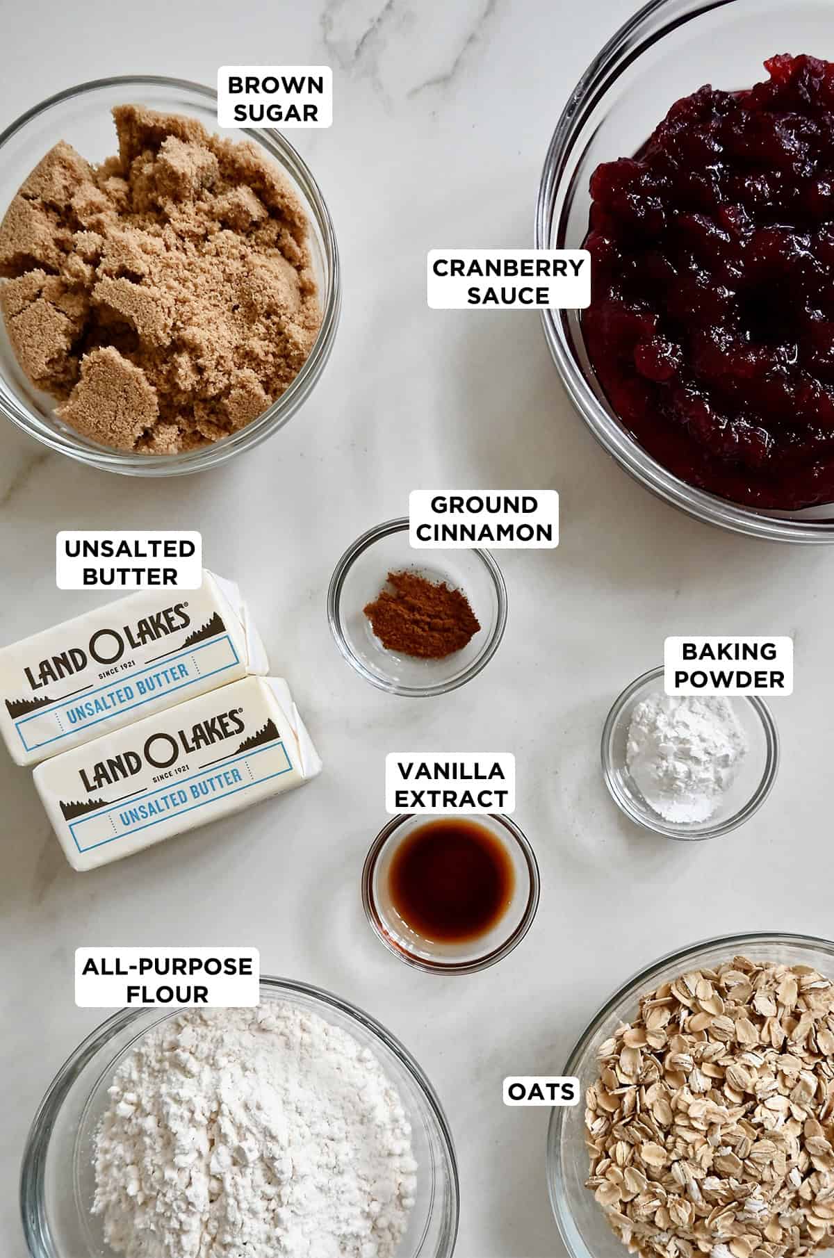 Various sizes of glass bowls containing the ingredients needed to make cranberry crumble pie bars, including cranberry sauce, ground cinnamon, butter, baking powder, vanilla extract, flour, rolled oats and brown sugar.