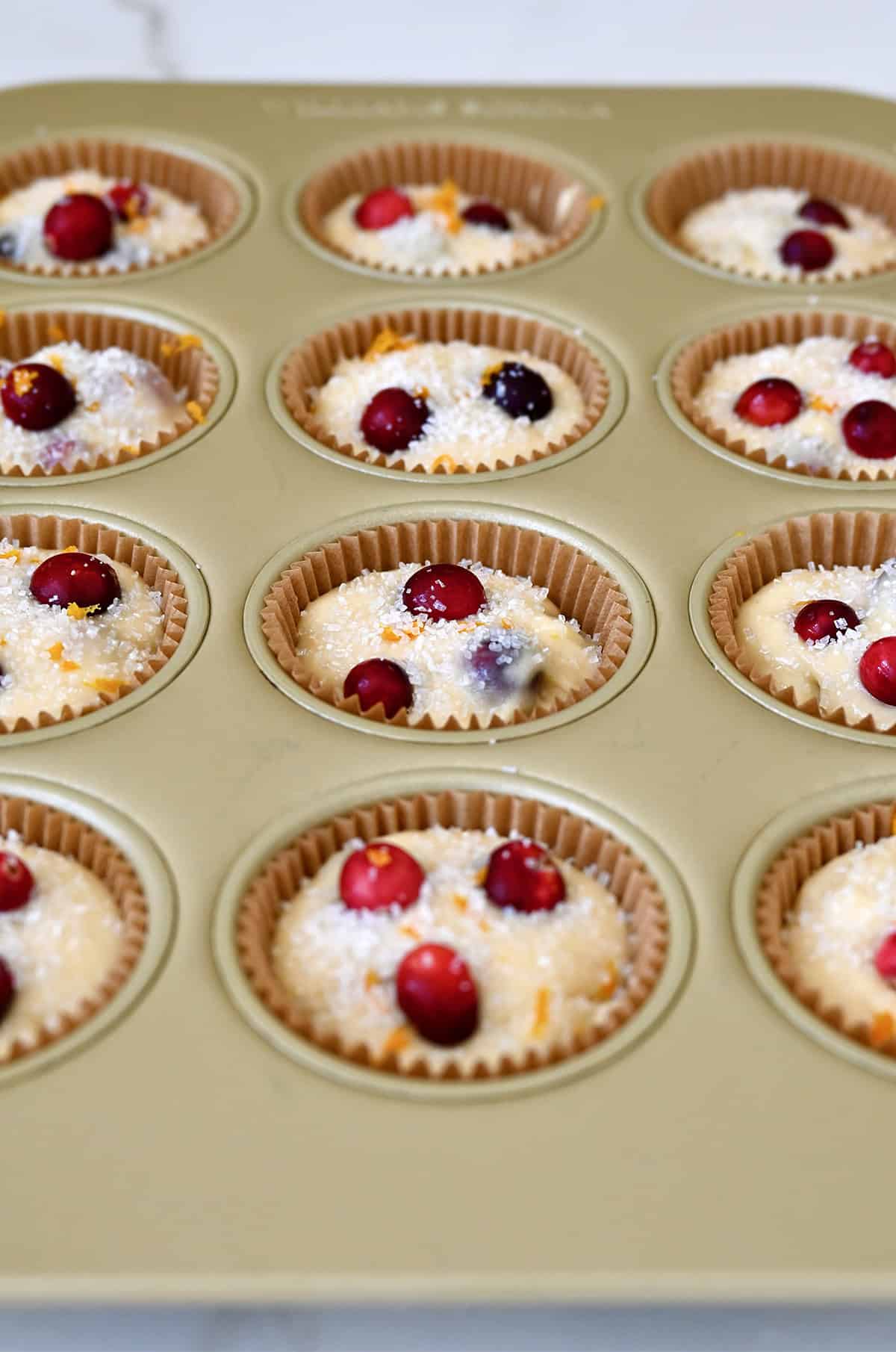 A standard muffin tin lined with paper liners filled with orange cranberry muffin batter, topped with sanding sugar.