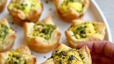 A hand holds a mini quiche with a puff pastry crust.