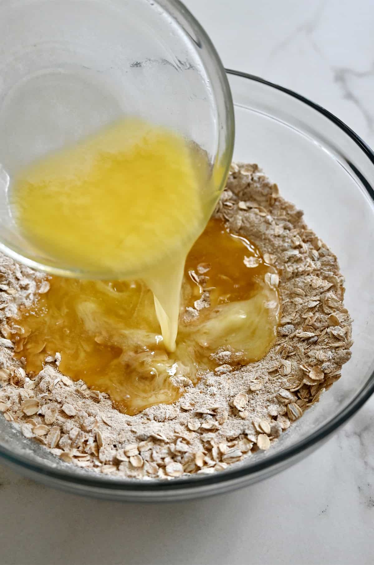Pouring melted butter from a small bowl into a larger bowl with an oat-flour-brown sugar mixture to make a crumb topping.