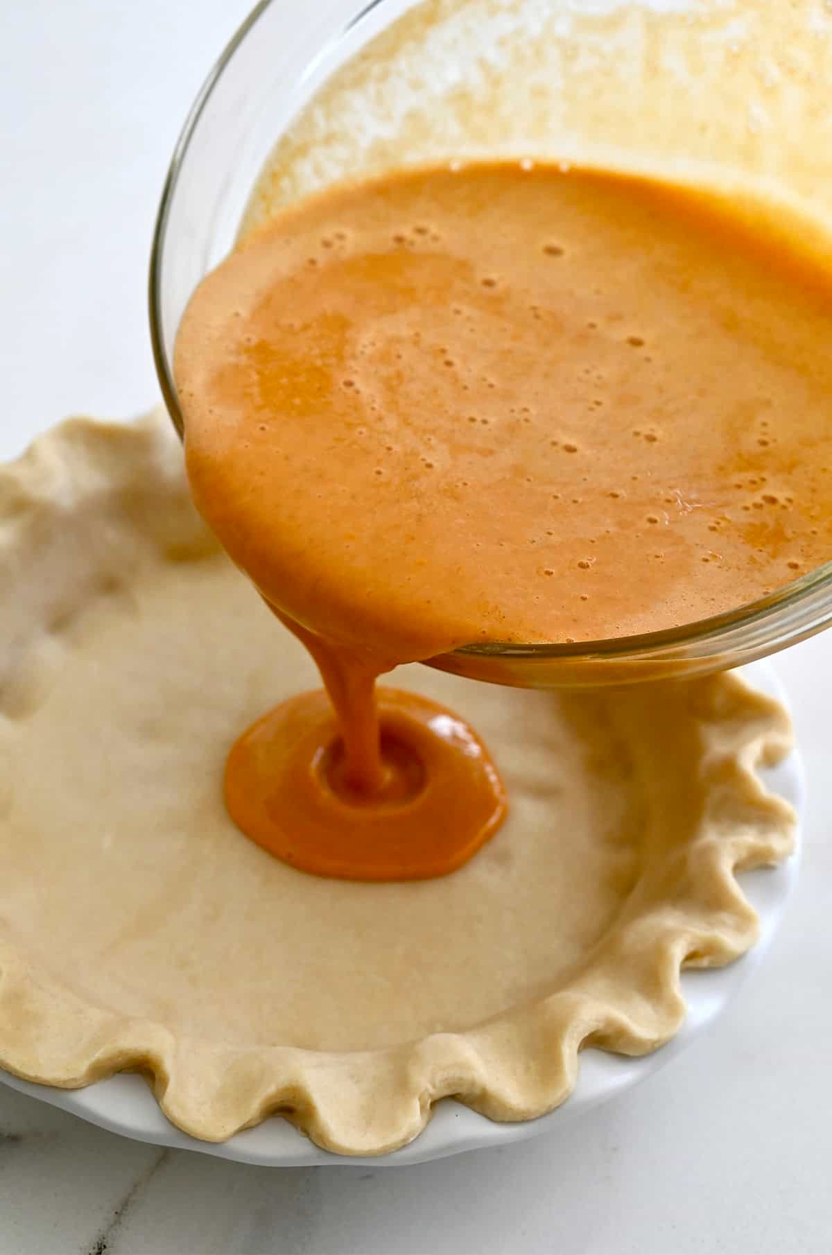 Pouring pumpkin pie filling from a glass bowl into a deep dish pie crust.