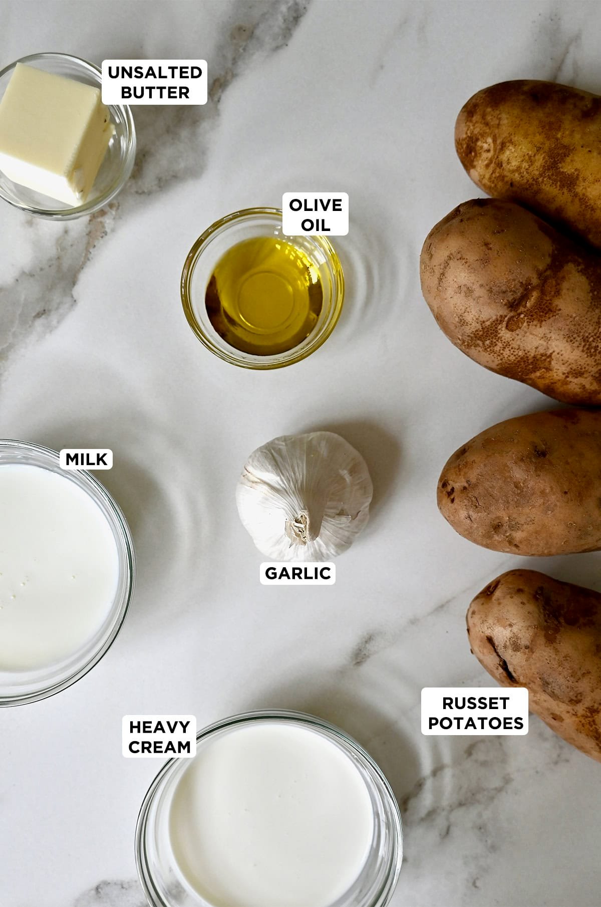 Four russet potatoes next to a head of garlic and bowls containing heavy cream, milk, butter and olive oil.