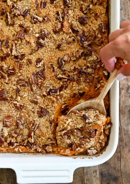 A hand holding a wooden spoon scoops a serving of creamy sweet potato casserole topped with golden brown pecan crumble from a white baking dish.