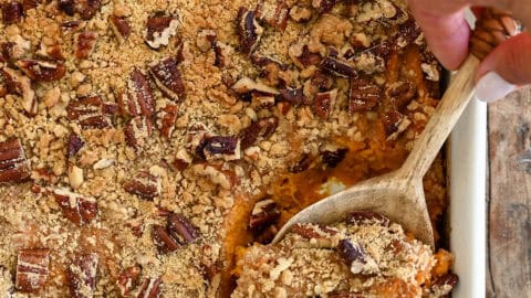 A hand holding a wooden spoon scoops a serving of creamy sweet potato casserole topped with golden brown pecan crumble from a white baking dish.