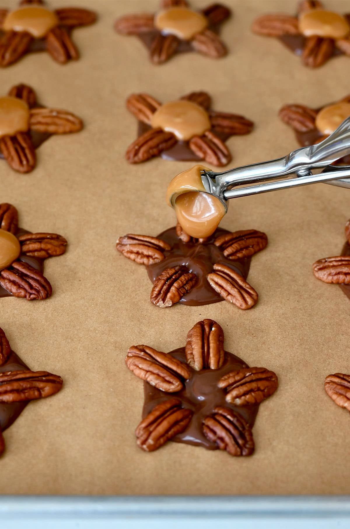 Spooning melted caramel on top of melted chocolate and pecan clusters on a parchment paper-lined baking sheet.
