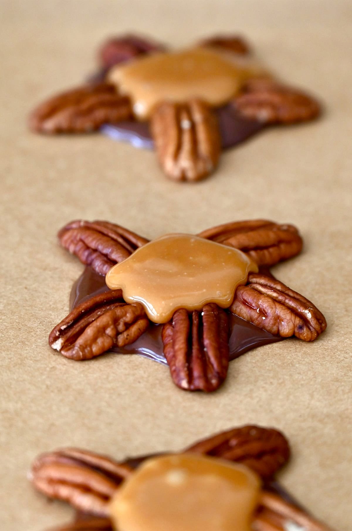 Spooning melted caramel on top of melted chocolate and pecan clusters on a parchment paper-lined baking sheet.