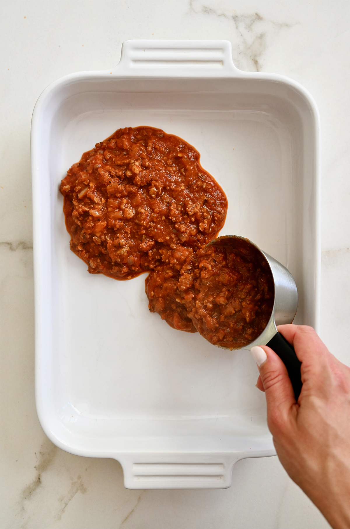 Adding meat marinara sauce to the bottom of a casserole dish for the first layer of lasagna.