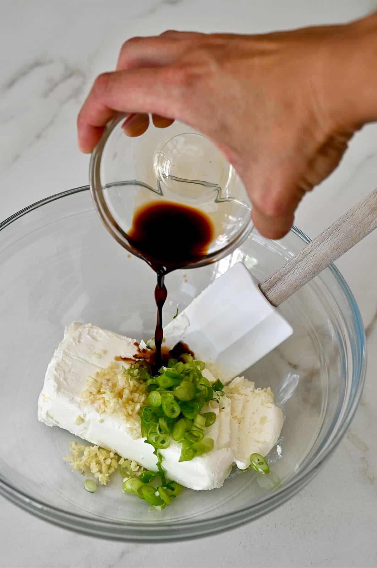 Adding soy sauce to a bowl containing cream cheese, sliced scallions and minced garlic.