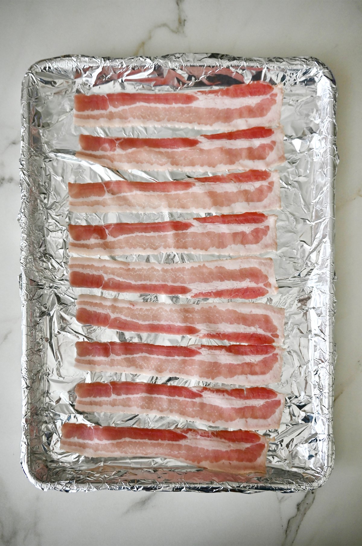 Uncooked bacon on an aluminum foil-lined baking sheet.