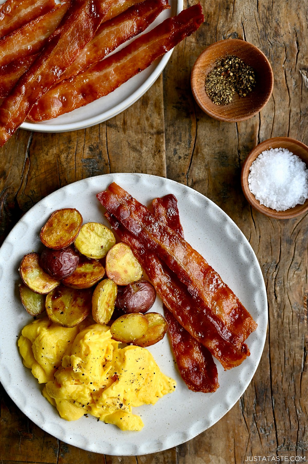Strips of crispy bacon on a plate with scrambled eggs and breakfast potatoes.