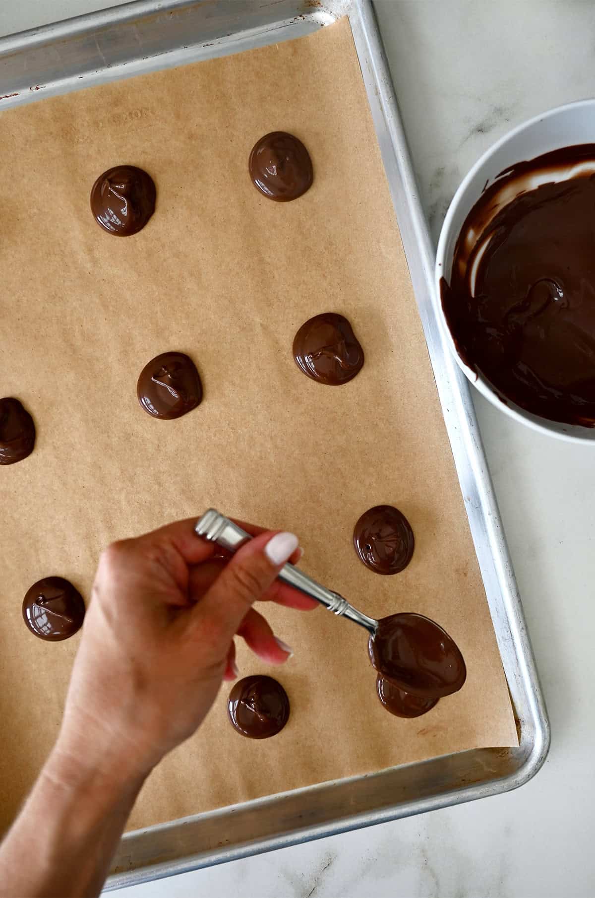 Creating the bottom layer of chocolate turtle candies by spooning melted chocolate into 1-tablespoon portions on a parchment paper-lined baking sheet.