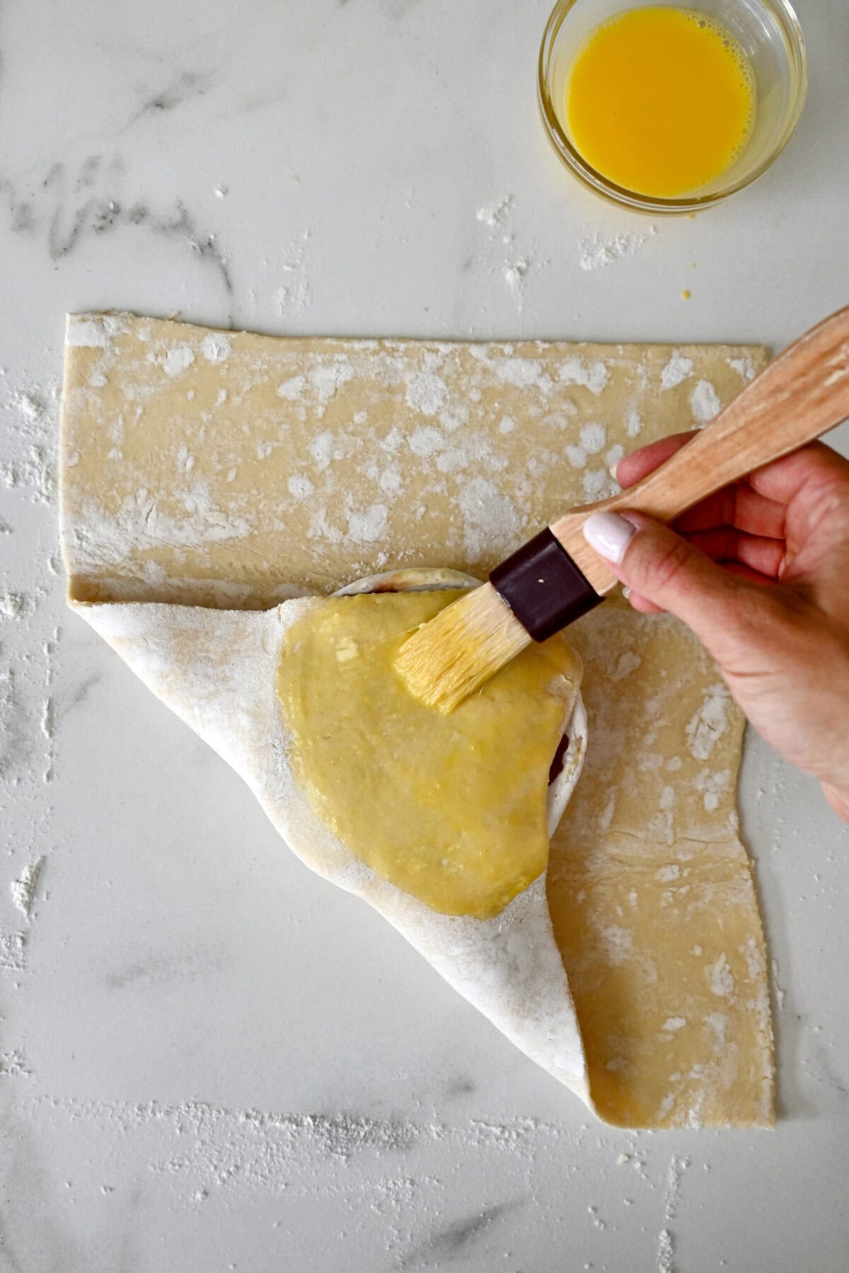 Brushing egg wash on top of puff pastry folded over a wheel of Brie topped with jam.