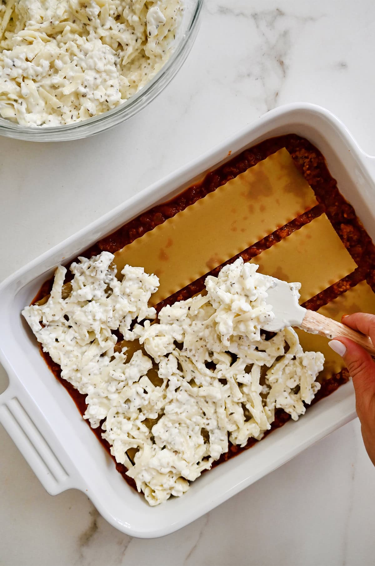 Adding the cheese layer atop lasagna noodles and meat marinara sauce in a casserole dish.