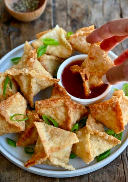 Crispy homemade crab Rangoon on a serving plate with one being dipped into sweet and sour sauce.