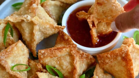 Crispy homemade crab Rangoon on a serving plate with one being dipped into sweet and sour sauce.