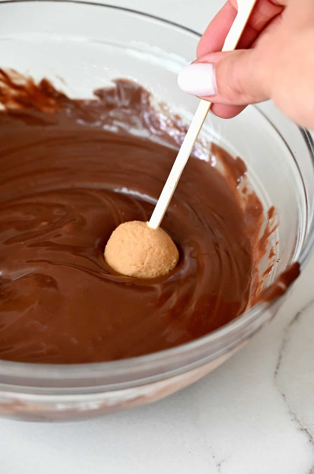 A peanut butter ball speared with a toothpick being dipped into melted chocolate.