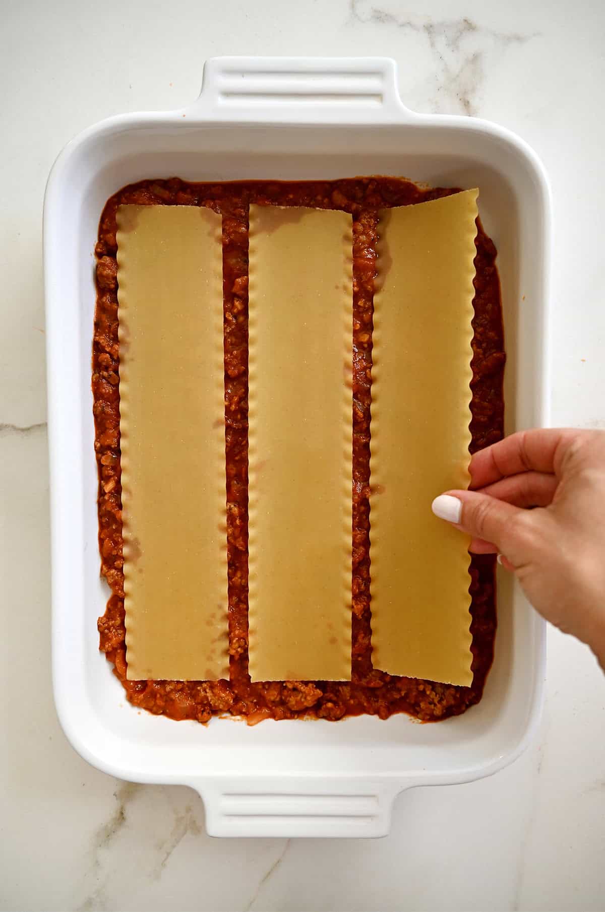 Adding three lasagna noodles atop a meat marinara sauce in a casserole dish.