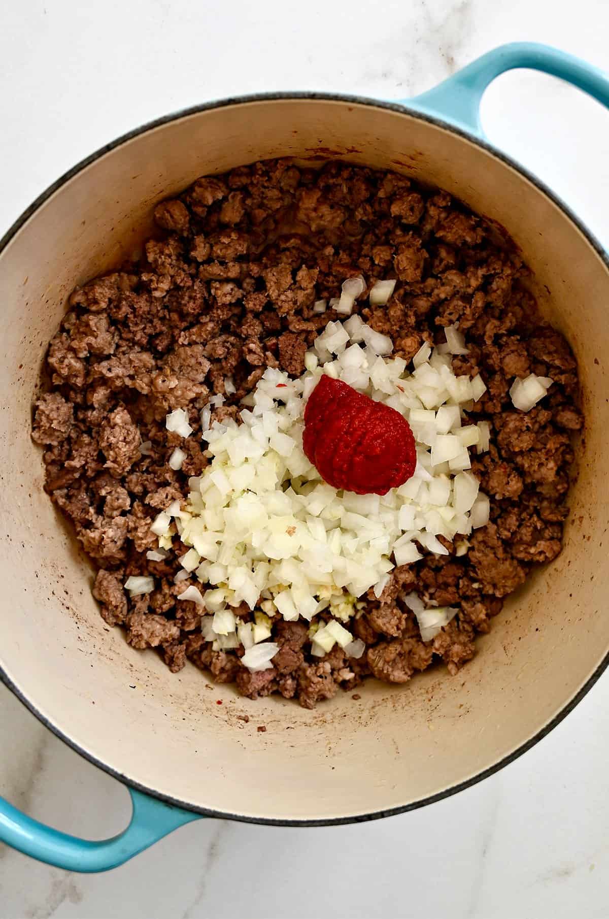 Browned ground beef and Italian sausage, topped with diced onion, minced garlic and tomato paste in a large pot.