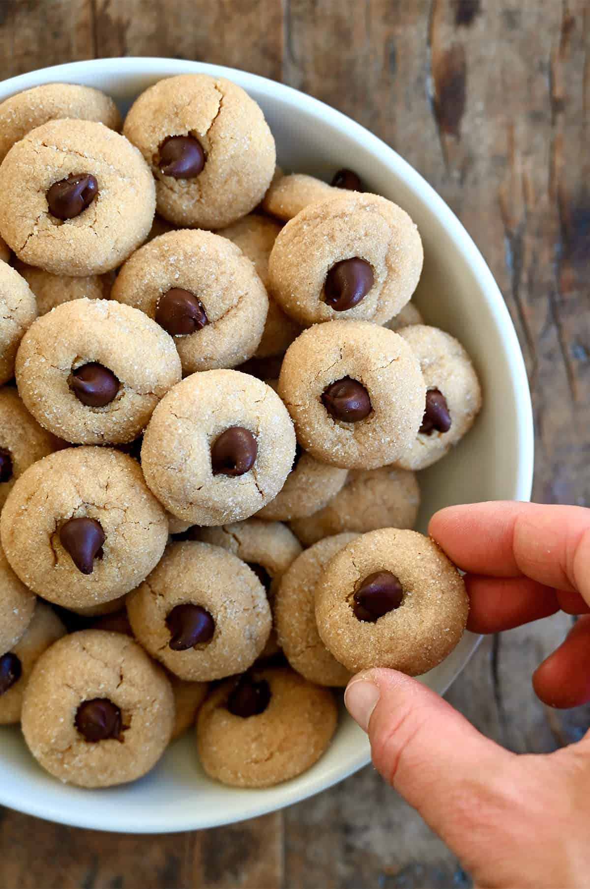 A hand reaching for a mini peanut butter blossom cookie in a bowl filled with mini peanut butter blossoms.