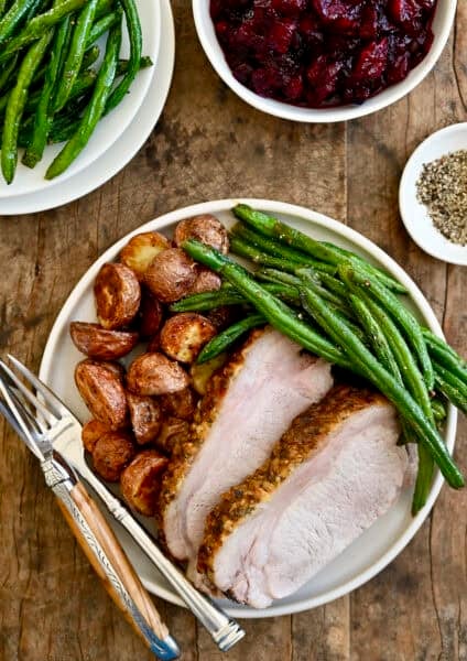 Two slices of herb-crusted pork loin roast on a dinner plate with roasted green beans and potatoes.