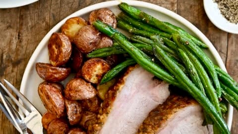 Two slices of herb-crusted pork loin roast on a dinner plate with roasted green beans and potatoes.