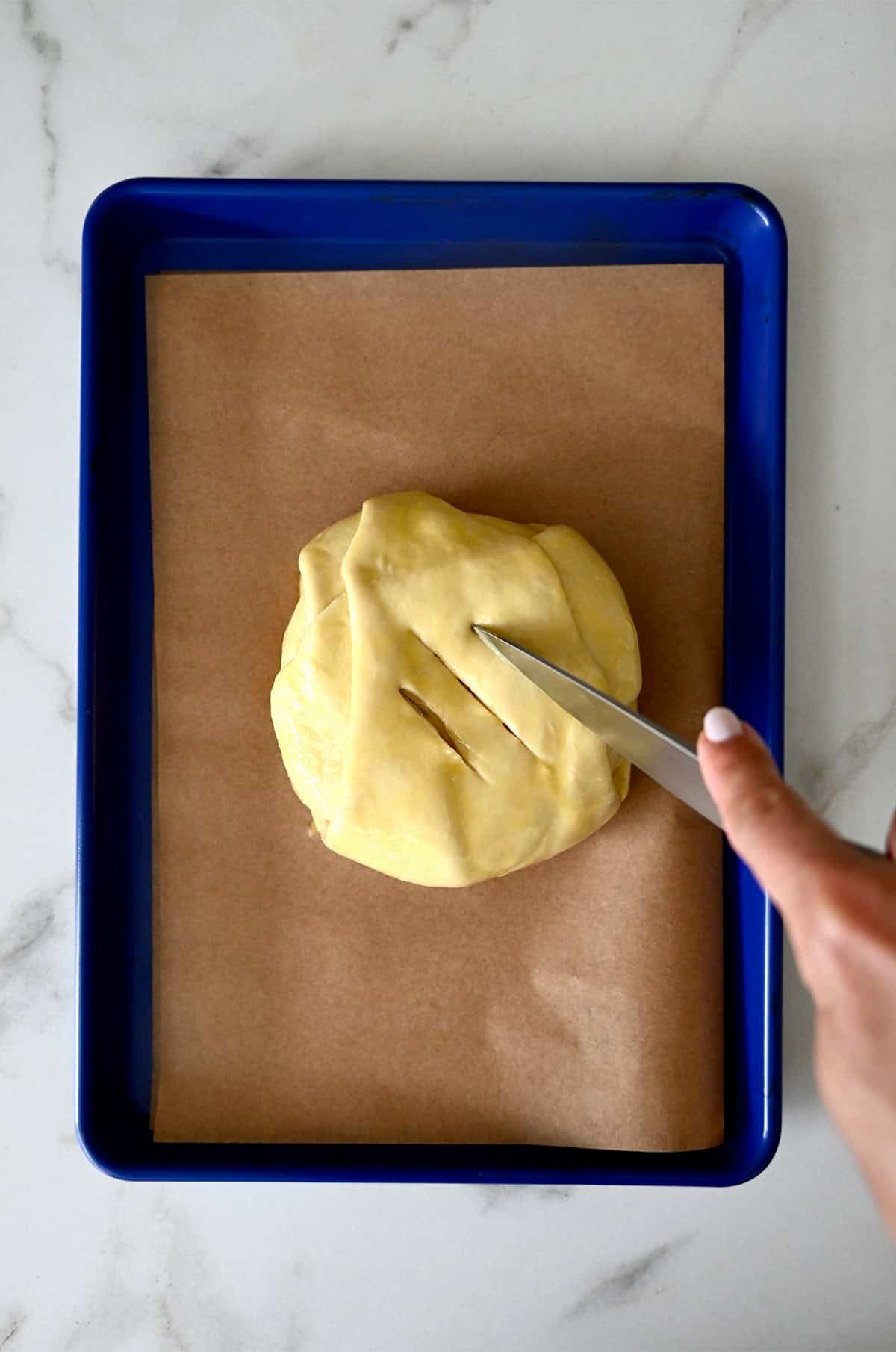 Brie cheese wrapped in puff pastry on a parchment paper-lined baking sheet. Three slits are being cut into the top of the pastry.