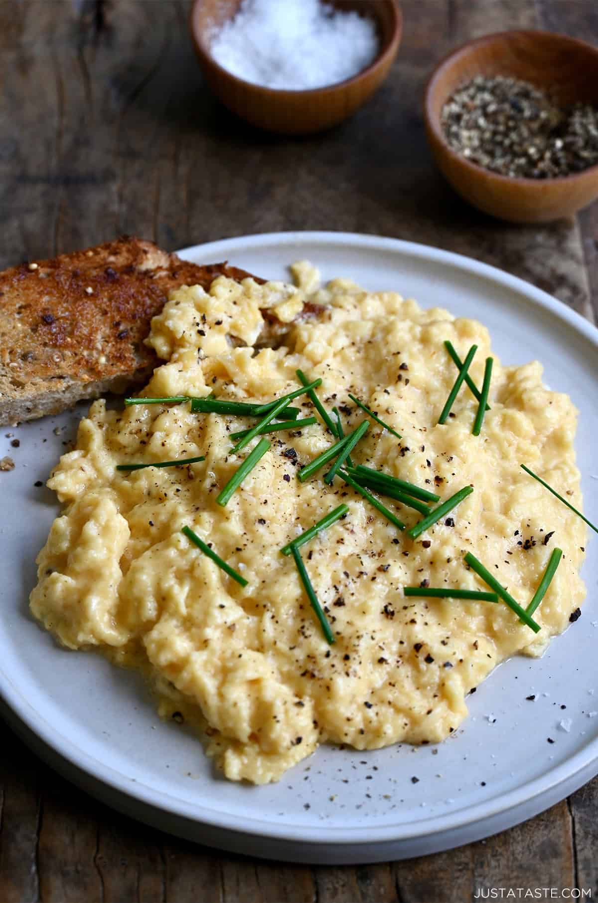 Soft scrambled eggs with freshly cracked black pepper, large flake sea salt and chives on a plate with a slice of toast.