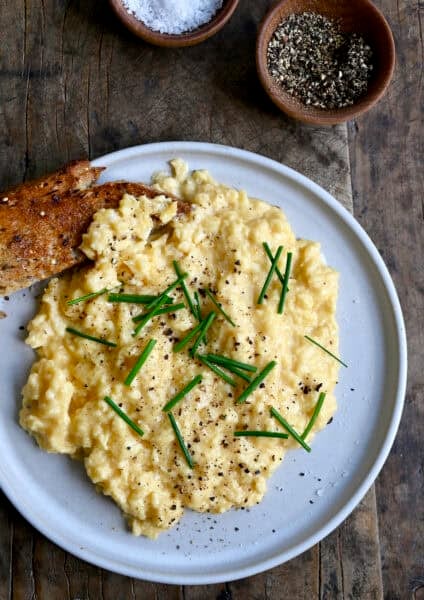 Soft scrambled eggs topped with salt, pepper and fresh chives on a plate with sourdough toast.