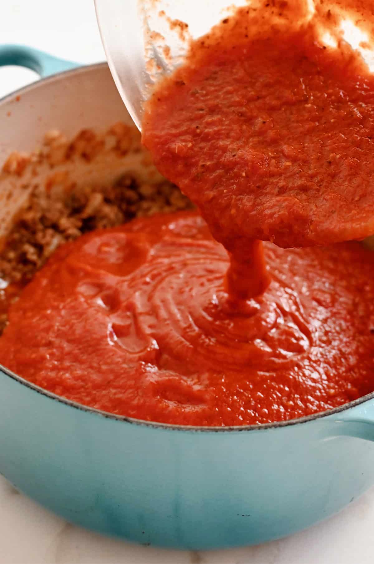 Adding tomato sauce to a pot with cooked ground meat to make sauce for lasagna.